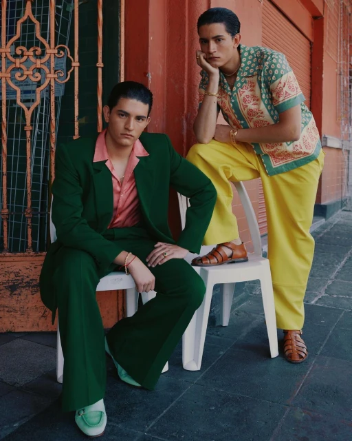 Two adult men in stylish 1970s-inspired fashion seated outdoors, one in a green suit and loafers, the other in printed top and yellow pants.