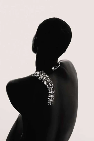 Back view of a woman with a shaved head and dark skin, wearing a silver necklace and a large, ornate jeweled shoulder piece.