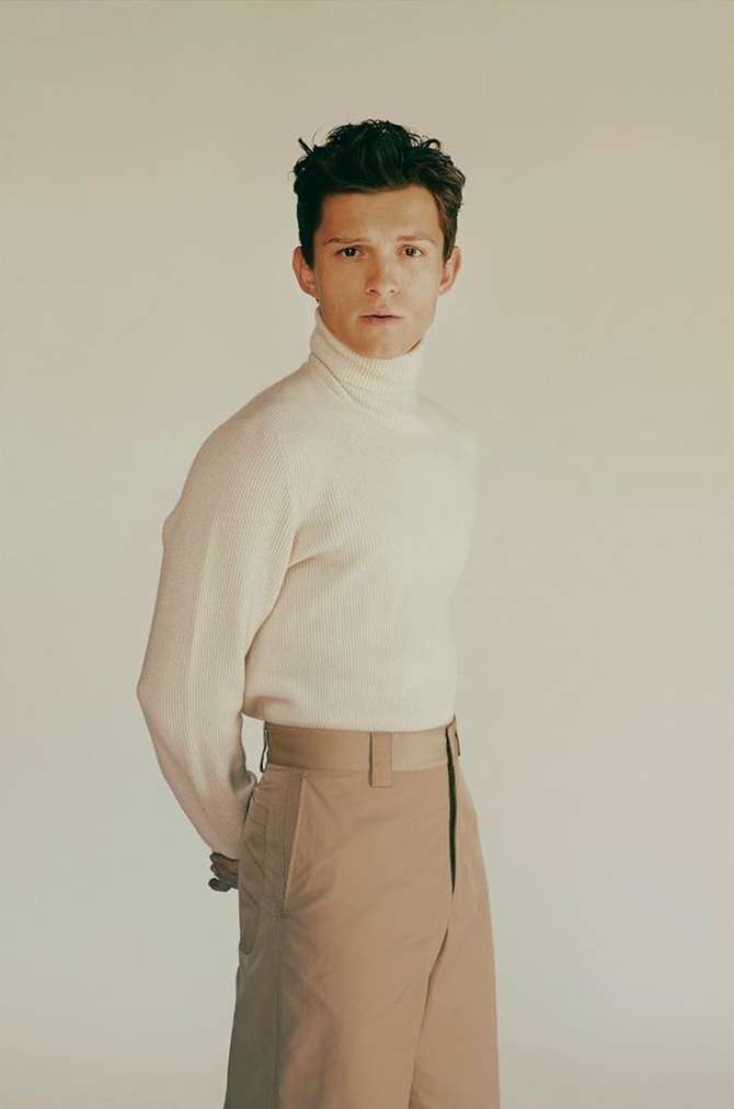 Tom Holland in a cream turtleneck sweater and khaki trousers against a seamless neutral background