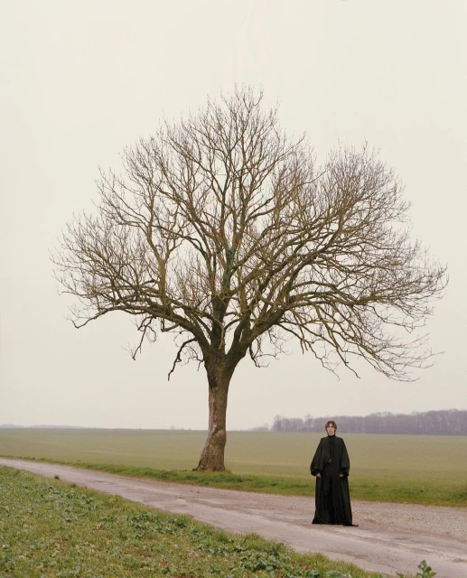 Woman in long black dress stands next to a large bare tree along a path in a rural landscape with fields and muted grey sky.