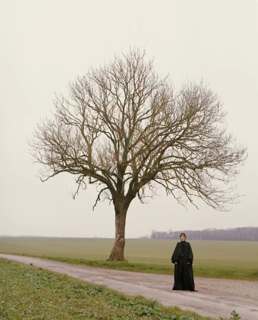 Woman in long black dress stands next to a large bare tree along a path in a rural landscape with fields and muted grey sky.
