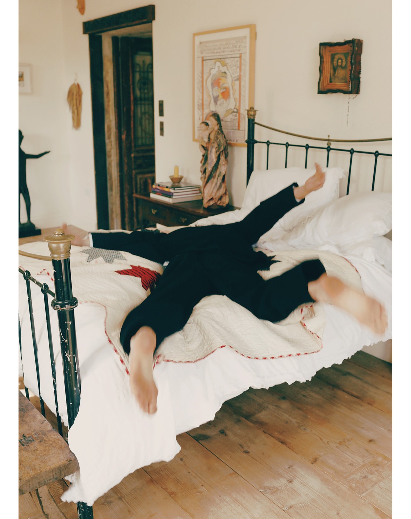 Person in suit lies on bed with white bedding. An expressive pose shows splayed arms and legs in an indoor bedroom setting.