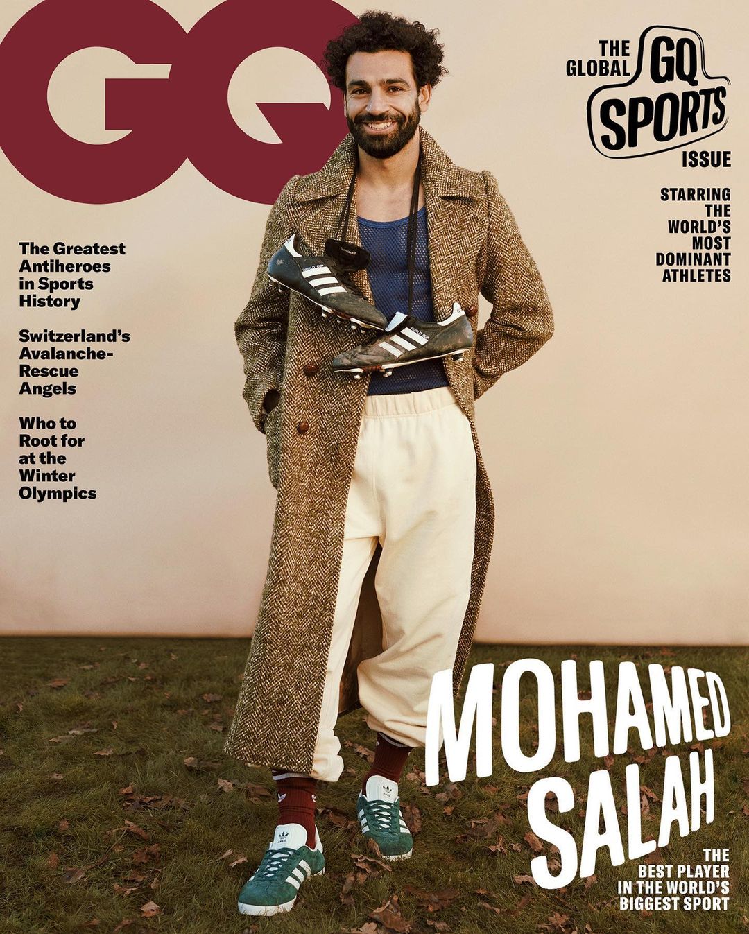 Mohamed Salah GQ cover featuring the athlete in a brown overcoat and sport pants holding soccer shoes, standing outdoors on grass.