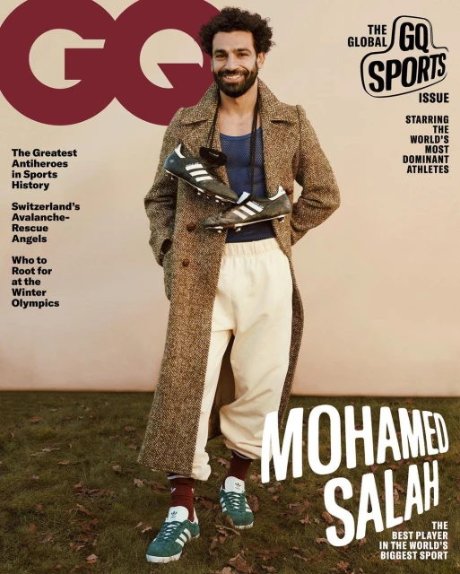 Mohamed Salah GQ cover featuring the athlete in a brown overcoat and sport pants holding soccer shoes, standing outdoors on grass.
