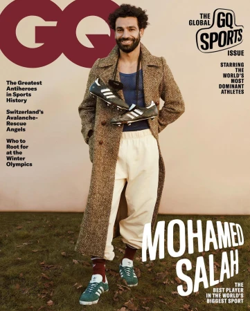 Mohamed Salah GQ cover featuring the athlete in a brown overcoat and sport pants holding soccer shoes, standing outdoors on grass.