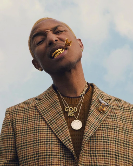 Portrait of Pharrell Williams wearing a brown plaid jacket, layers of gold necklaces, and gold grill teeth against a blue sky.