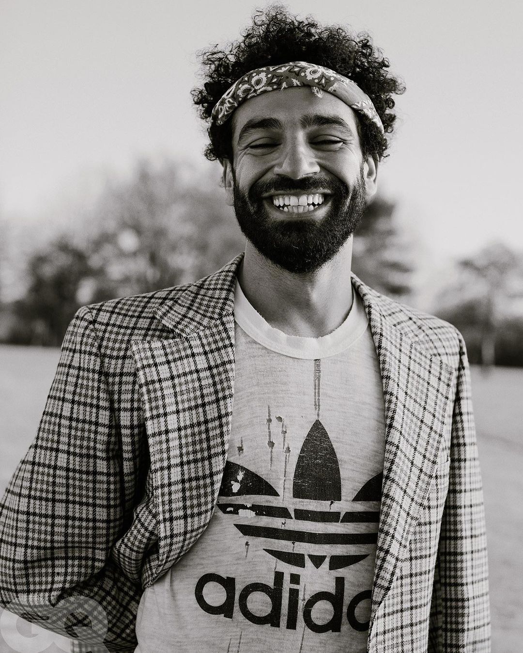 Black and white close-up of a man with curly hair, a bandana, and a printed T-shirt wearing a plaid blazer and smiling outdoors.