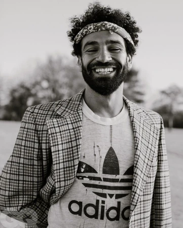 Black and white close-up of a man with curly hair, a bandana, and a printed T-shirt wearing a plaid blazer and smiling outdoors.