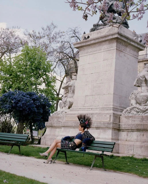 Woman in a patterned skirt suit and feather headpiece sits on a bench near a stone monument in a sunny park setting.