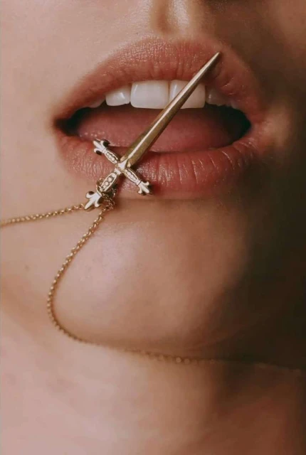 Close-up of a person's mouth with white teeth and an open mouth, featuring a sword-shaped necklace resting on the bottom lip.