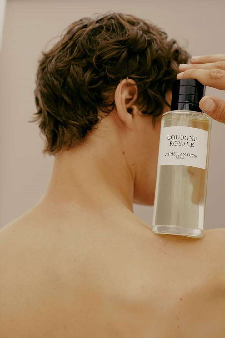 Adult male showing bare back and curly hair presenting a 'Cologne Royale' perfume bottle by Christian Dior Paris against a light wall.