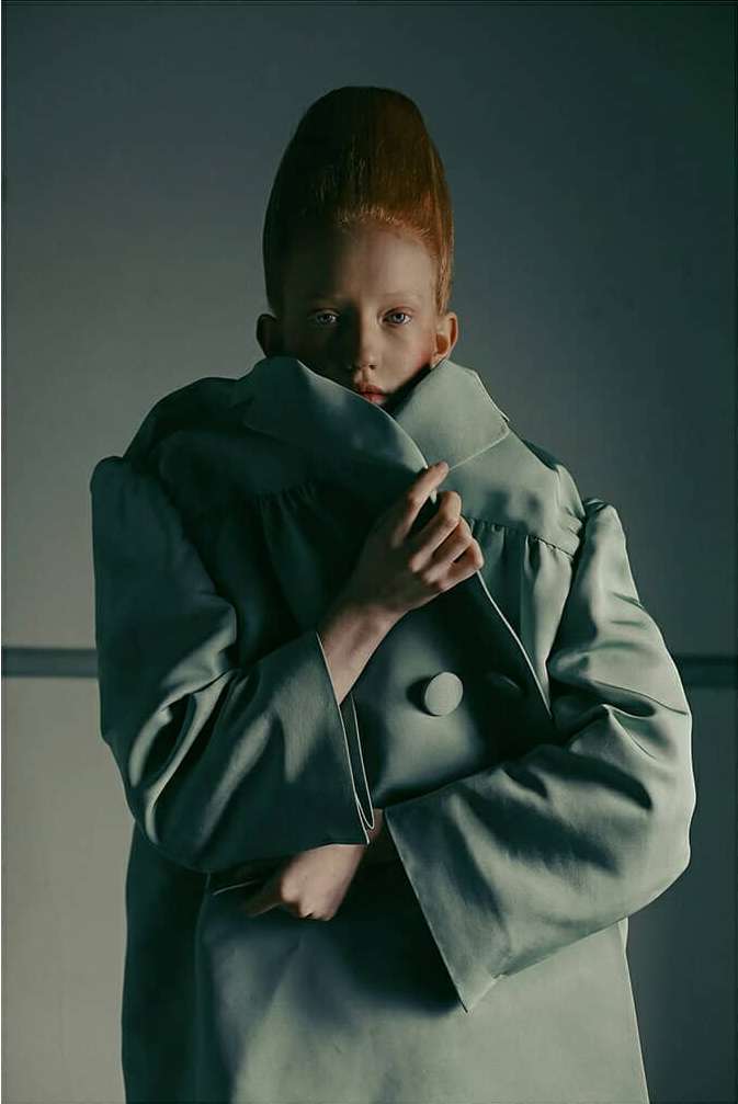 Woman with striking red hair and pale skin poses in a pale green, oversized button-down jacket in a modern studio fashion portrait.