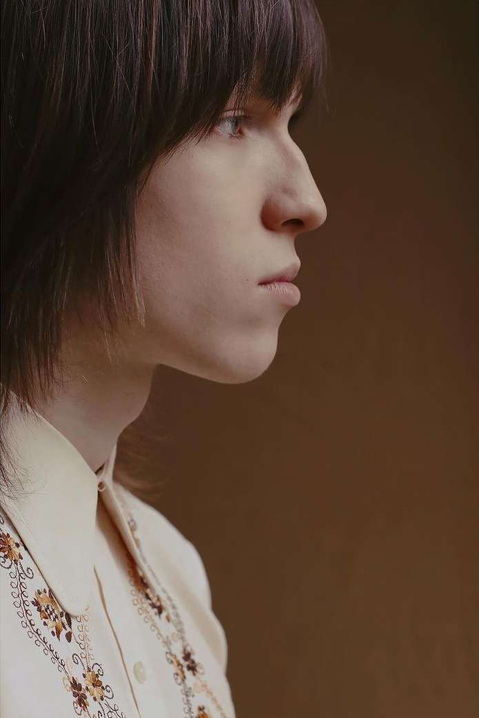 Portrait of a young adult with dark hair, posing in profile wearing an off-white shirt with an embroidered floral pattern against a tan backdrop.
