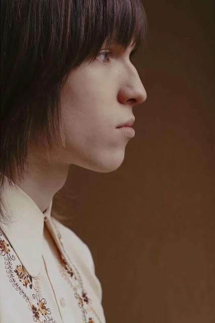 Portrait of a young adult with dark hair, posing in profile wearing an off-white shirt with an embroidered floral pattern against a tan backdrop.