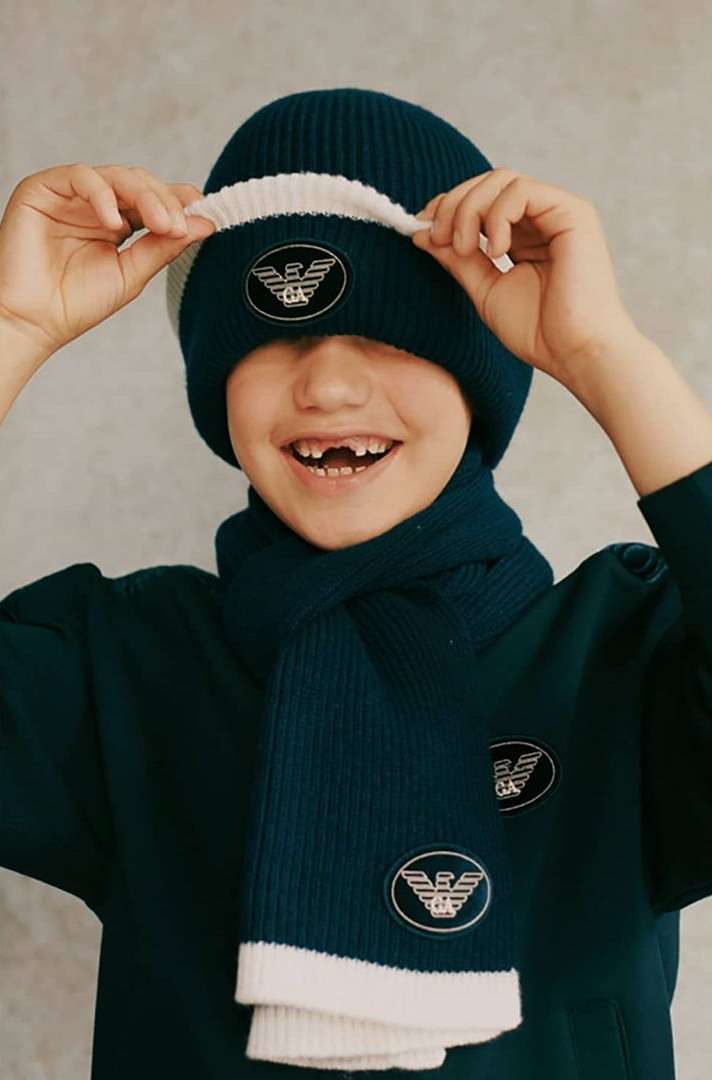 Child poses in blue knit hat and scarf with logo, looking at camera, against neutral backdrop, showcasing youth fashion.