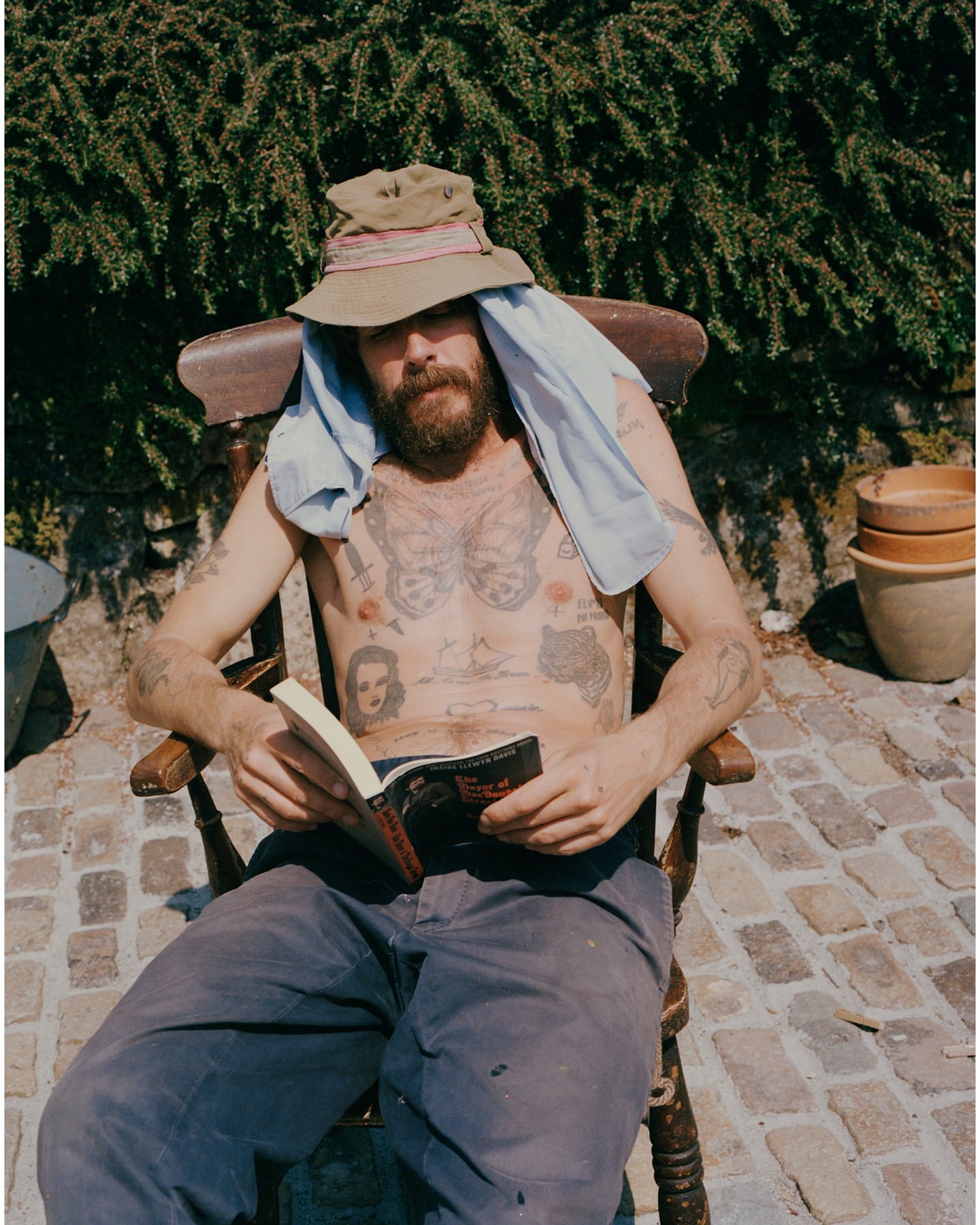 Man with beard and tattoos sits in a wooden chair outside, reading a book under bright sunlight, wearing a hat and a towel over his shoulders.