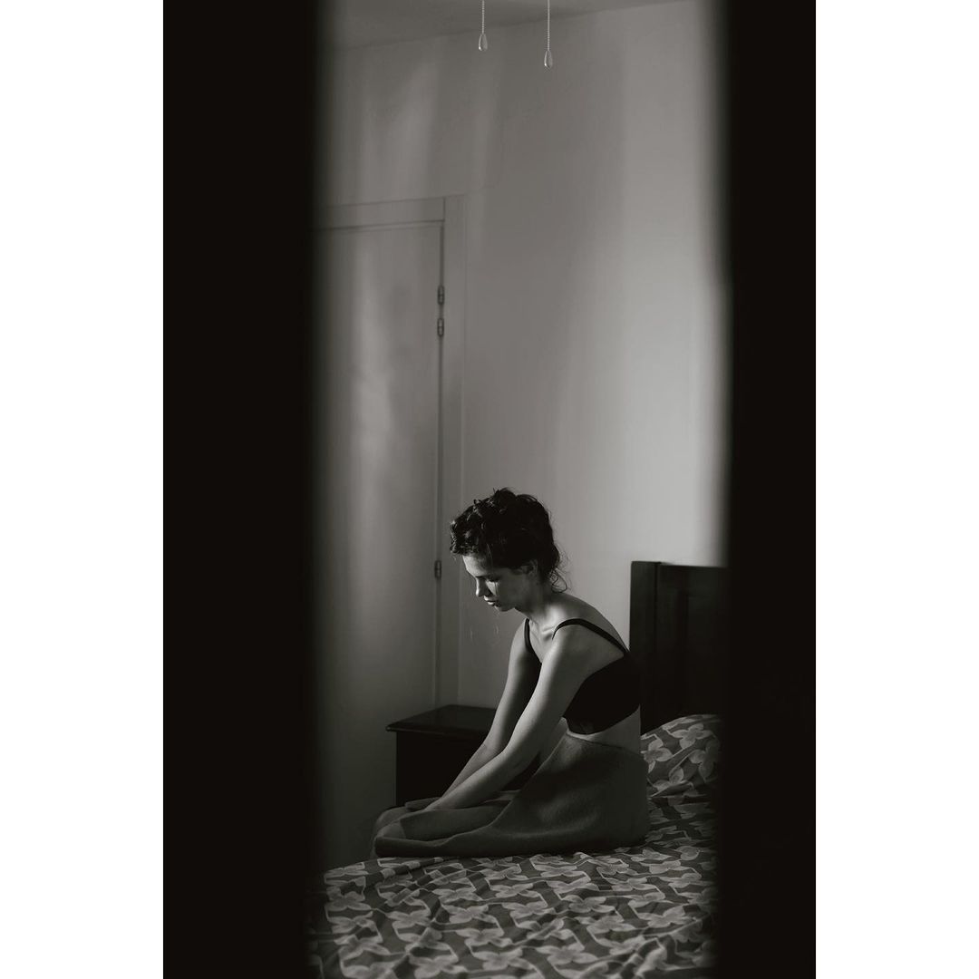 Woman sitting on a patterned bed in a bedroom viewed through a vertical gap by Fanny Latour-lambert