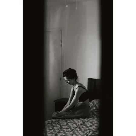 Woman sitting on a patterned bed in a bedroom viewed through a vertical gap by Fanny Latour-lambert
