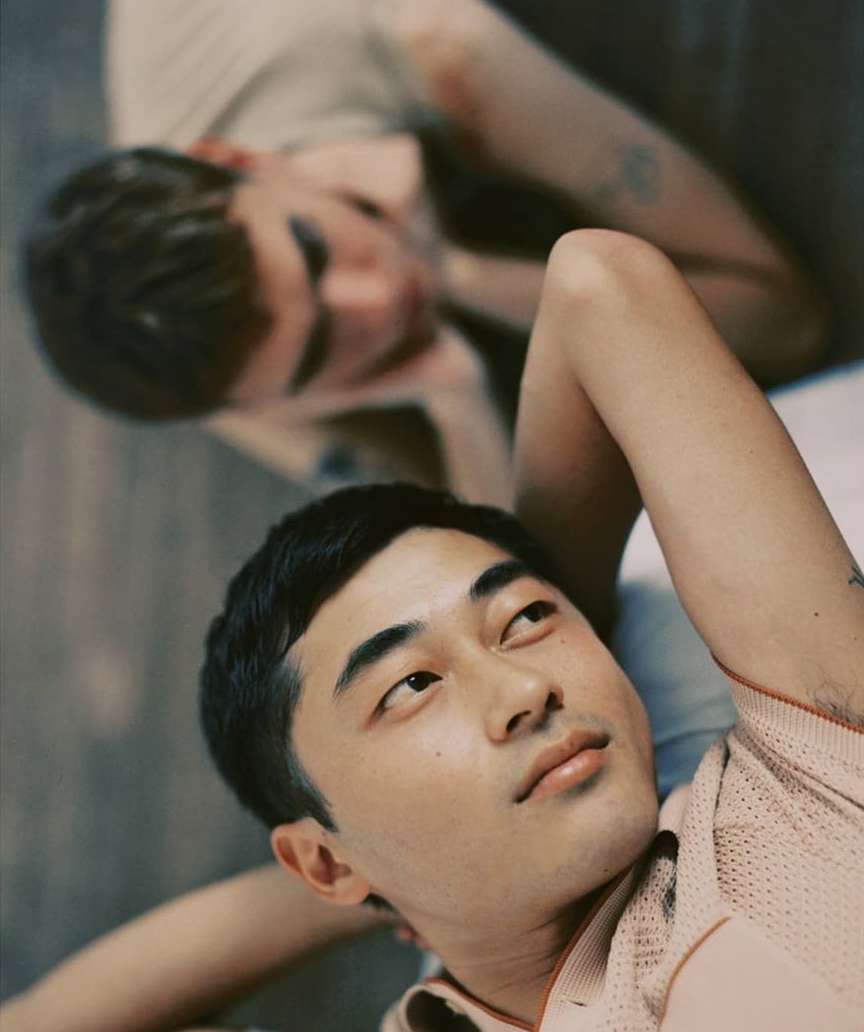 Close-up portrait of two young adult males indoors; one with short dark hair looking up, the other out of focus behind him.