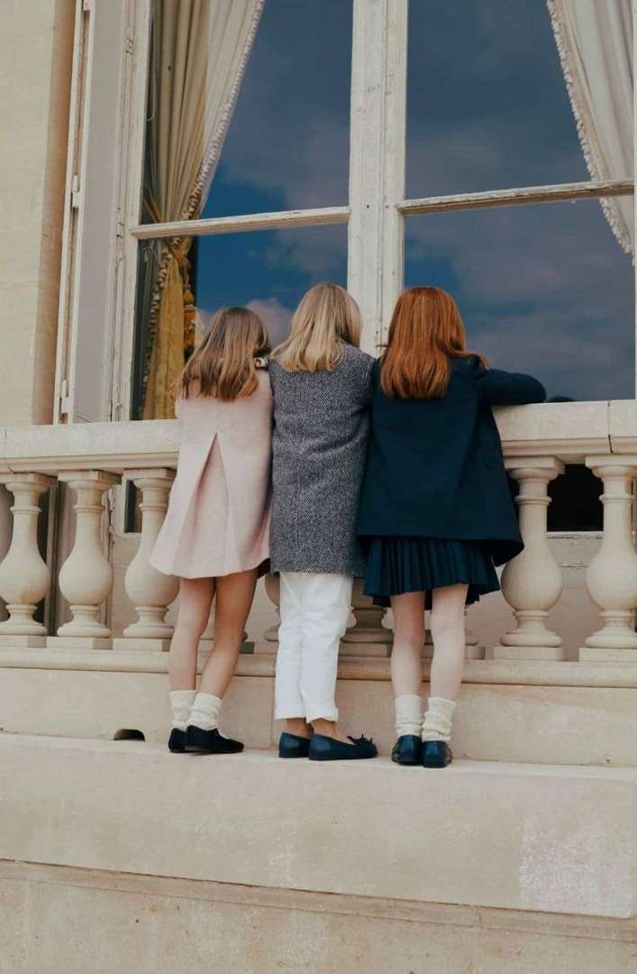 Three young girls stand shoulder-to-shoulder looking out a window of a building with cream-colored walls and white trim and balustrade.