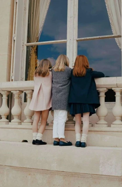 Three young girls stand shoulder-to-shoulder looking out a window of a building with cream-colored walls and white trim and balustrade.