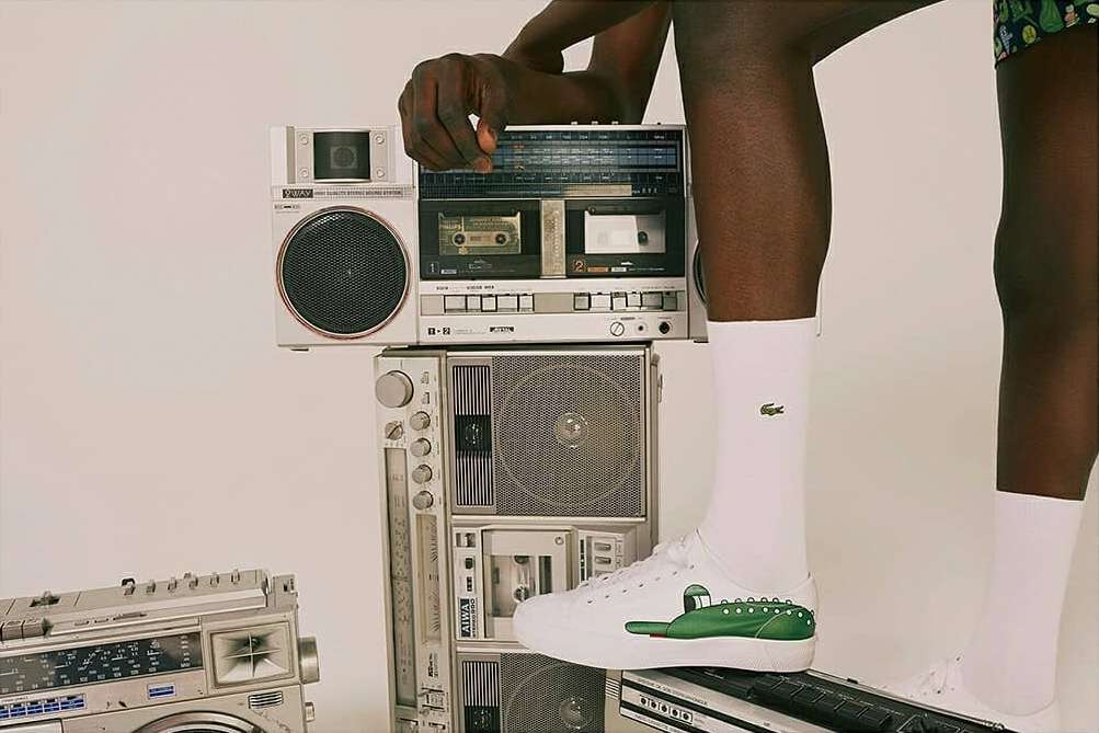 Close-up of a stack of vintage silver boomboxes with a male model wearing white Lacoste sneakers and socks standing on top.