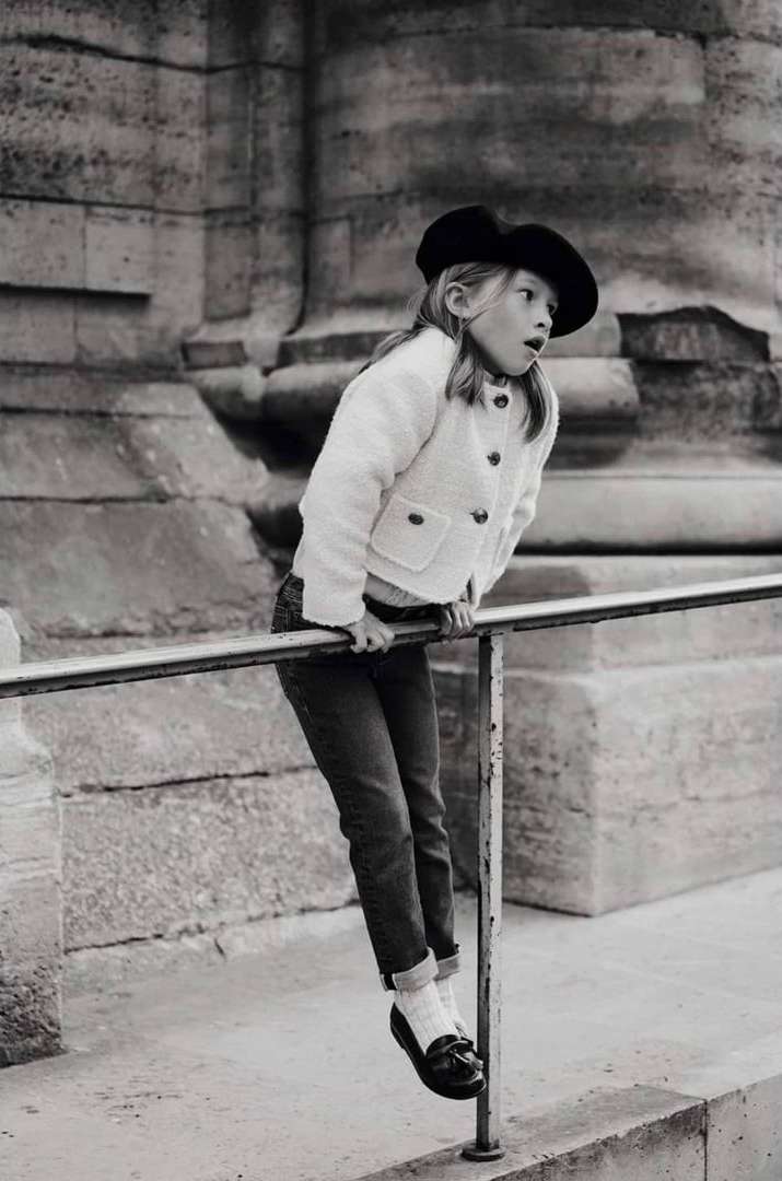 Young child in a beret and textured jacket hanging playfully from a metal railing outdoors by classical architecture.