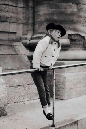 Young child in a beret and textured jacket hanging playfully from a metal railing outdoors by classical architecture.