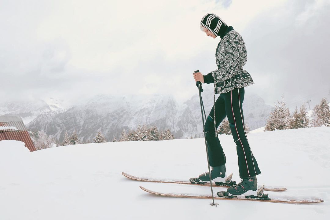 A woman in winter fashion skis down a snowy slope with mountains in the background on a cloudy day, holding ski poles.