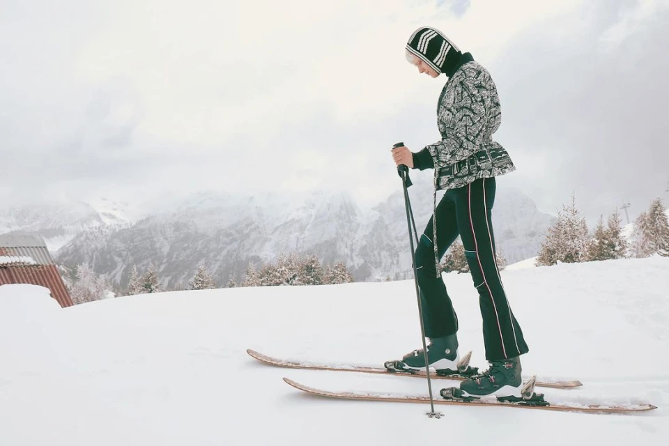 A woman in winter fashion skis down a snowy slope with mountains in the background on a cloudy day, holding ski poles.