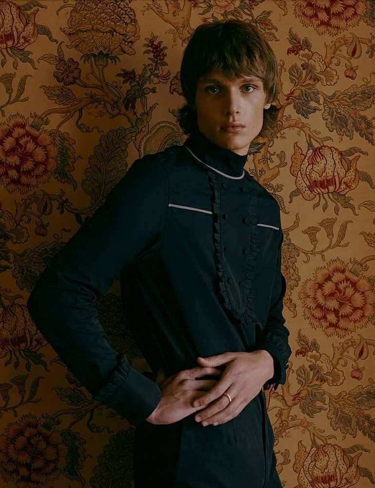 Young adult male model poses in a long-sleeved black shirt against a backdrop of antique floral patterned wallpaper.