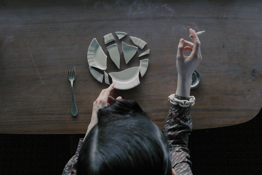 Overhead view of a dark wooden table with a woman holding a cigarette, a shattered plate, and a fork alongside muted lighting.