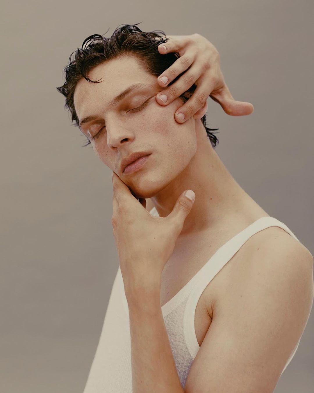 Portrait of a male fashion model with closed eyes. Another person's hands are gently touching his face in a studio setting.