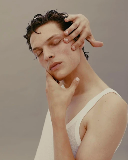 Portrait of a male fashion model with closed eyes. Another person's hands are gently touching his face in a studio setting.