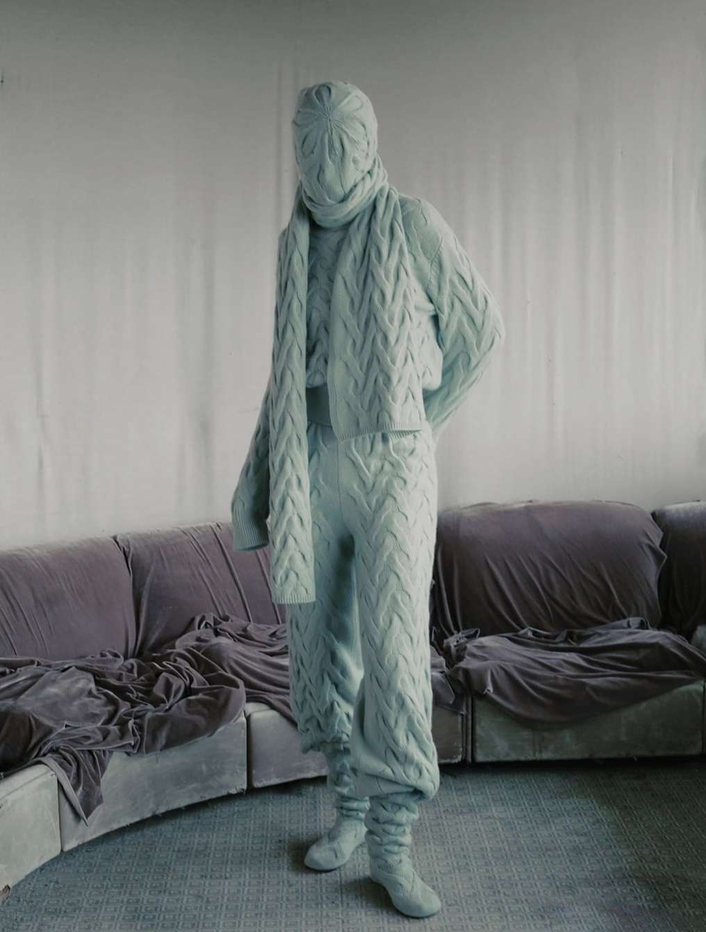 Person in knitted full-body suit stands in interior room with mauve seating. Pale color palette with soft light and geometric knit patterns.