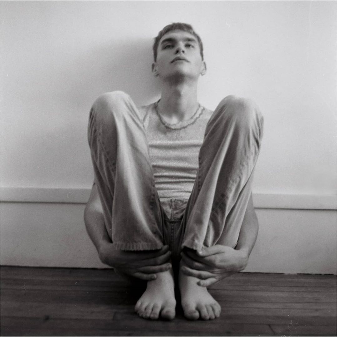 Monochrome portrait of a young adult male sitting with legs drawn up, casual wear, bare feet, against a white wall and wood floor.