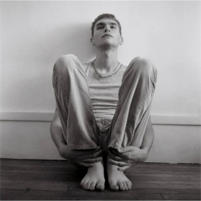 Monochrome portrait of a young adult male sitting with legs drawn up, casual wear, bare feet, against a white wall and wood floor.