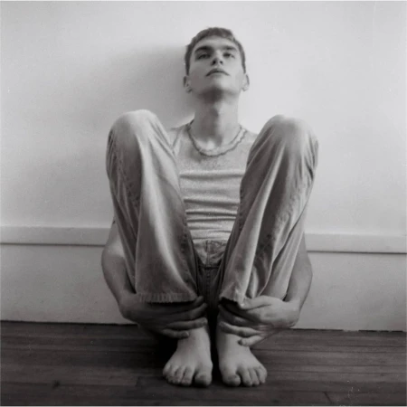 Monochrome portrait of a young adult male sitting with legs drawn up, casual wear, bare feet, against a white wall and wood floor.