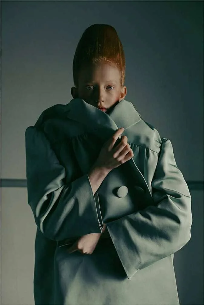 Model with sculpted red hair wearing a sage green oversized coat pulled high around neck by photographer Fanny Latour-lambert