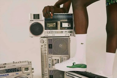 Person wearing white socks and sneakers standing beside stacked vintage boomboxes against a pale background
