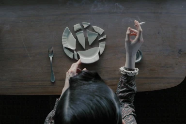 Top-down view of a person seated at a dark wooden table with broken ceramic plate shards, a fork, and a smoking cigarette.