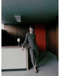 Model wearing a fitted grey jumpsuit and chunky necklace standing inside a wood-paneled interior by Fanny Latour-lambert