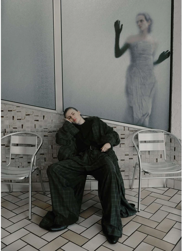 Woman in dark plaid suit seated on a metal chair against a tiled wall with a frosted glass window showing a figure silhouette.