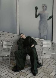Woman in dark plaid suit seated on a metal chair against a tiled wall with a frosted glass window showing a figure silhouette.