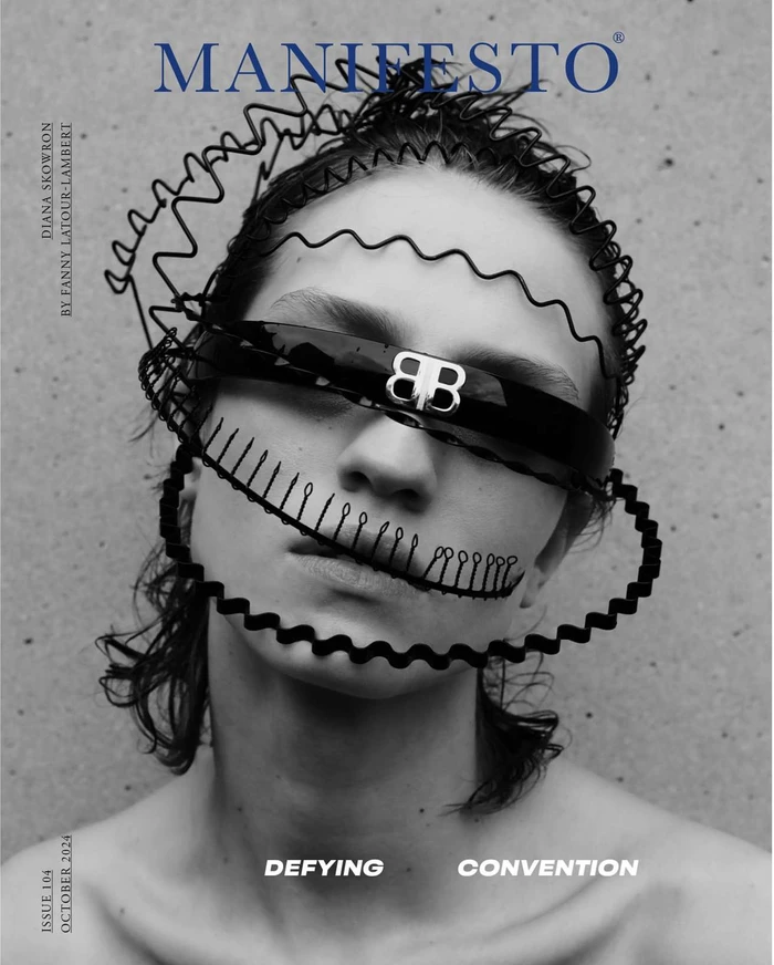 Close-up portrait of a model wearing sunglasses and a wire crown accessory against a concrete wall
