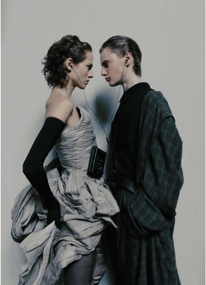Two women standing face to face sharing earphones while wearing contrasting grey gown and dark plaid coat by Fanny Latour-lambert