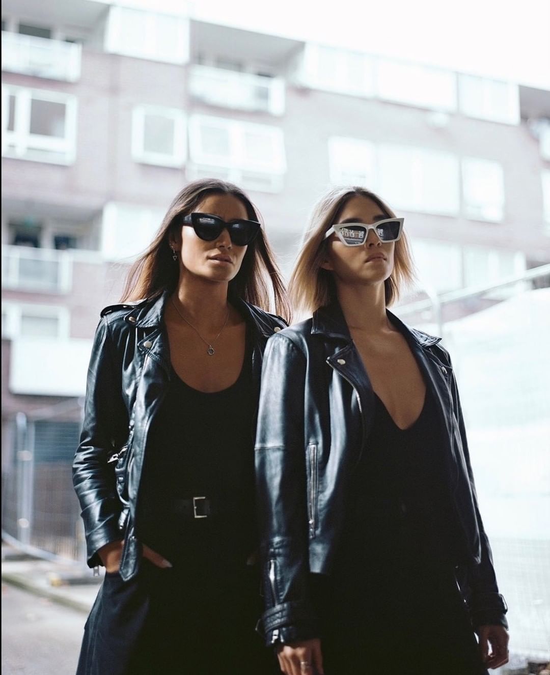 Two women in leather jackets and sunglasses pose outdoors in an urban environment. Fashion editorial featuring modern style and neutral colors.