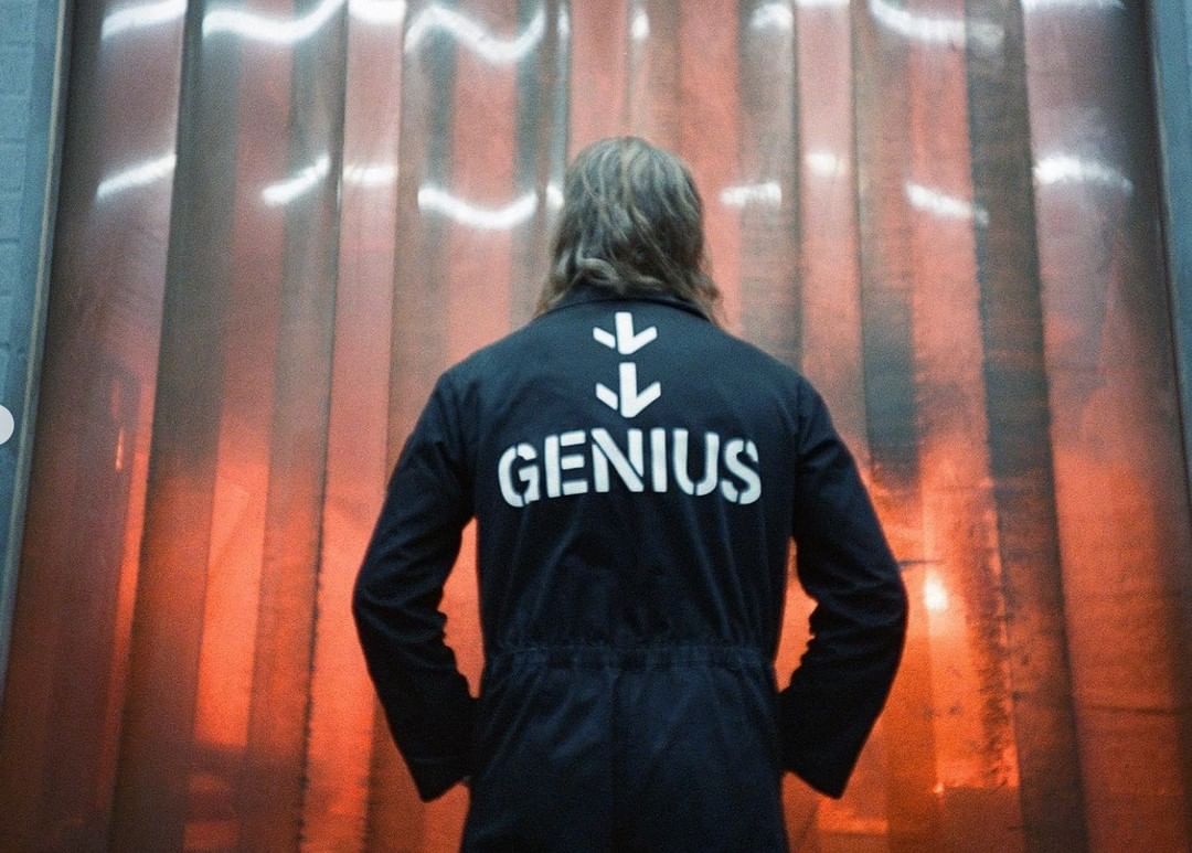 Rear view of person with long hair wearing a navy jumpsuit with the word "GENIUS" on the back standing indoors.