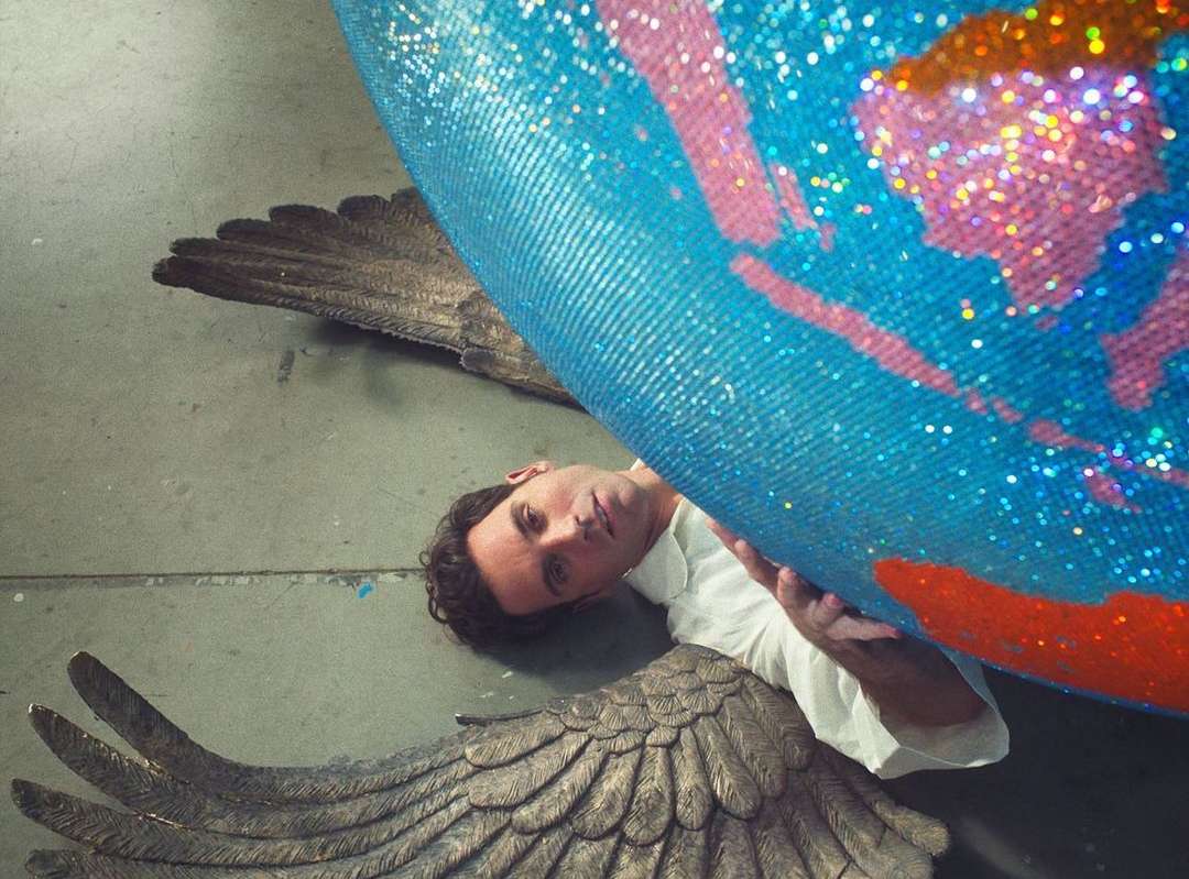 Artistic overhead shot of a man lying on the floor, supporting a glittery, painted globe adorned with wings, in an indoor setting.