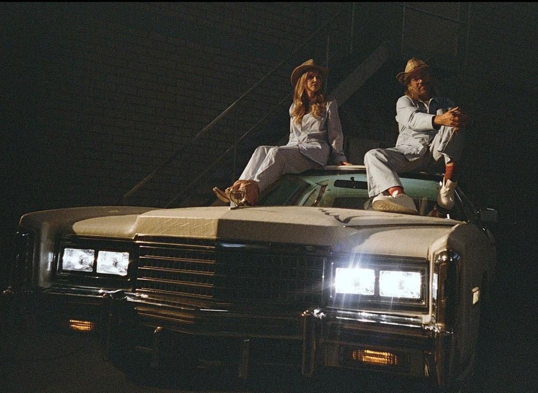 A man and woman in matching light blue suits and hats sit on a vintage Cadillac car against a dark brick wall background.